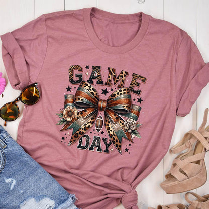 May include: A pink t-shirt with a graphic of a leopard print bow with glitter and the words "GAME DAY" in black letters.