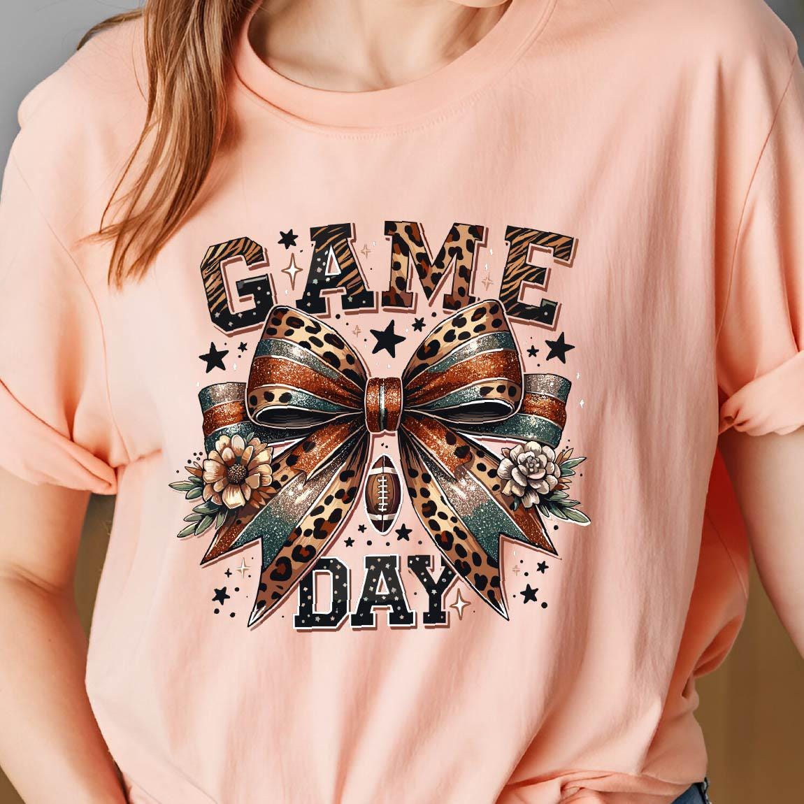 May include: A pink t-shirt with a graphic of a leopard print bow with glitter and the words "GAME DAY" in black.
