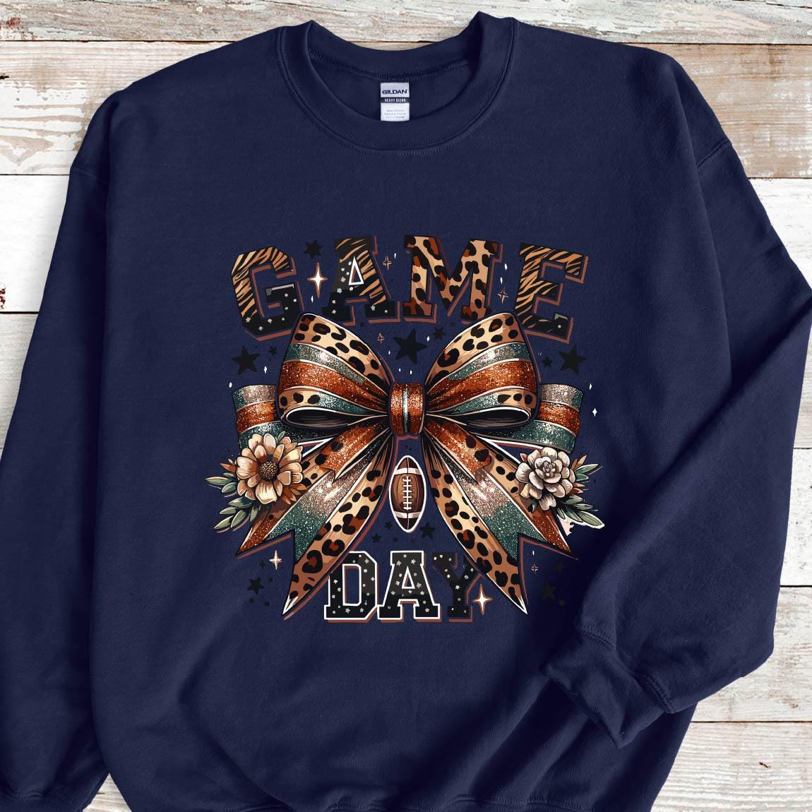 May include: Navy blue sweatshirt with a graphic of a leopard print bow with gold glitter accents and the text "GAME DAY" in a black and white font.