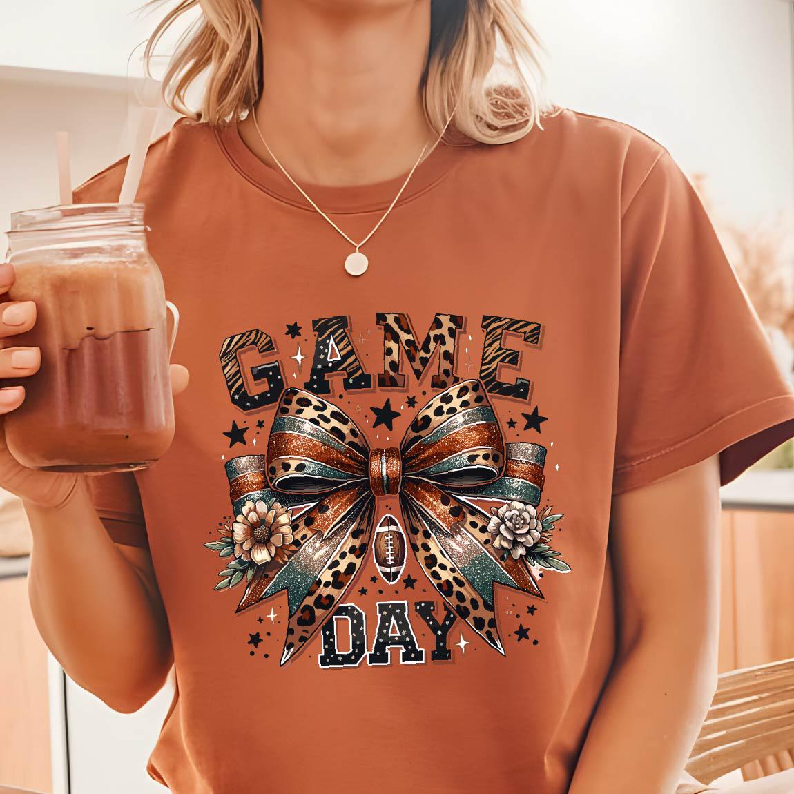 May include: A rust-colored t-shirt featuring a graphic print with the text GAME DAY in block letters. The design includes a large central bow with leopard print and teal accents, a small football icon, floral elements, and scattered stars.