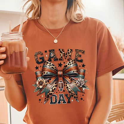 May include: A rust-colored t-shirt featuring a graphic print with the text GAME DAY in block letters. The design includes a large central bow with leopard print and teal accents, a small football icon, floral elements, and scattered stars.