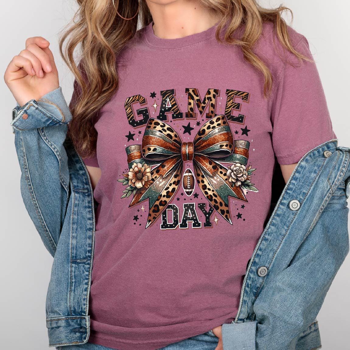 May include: A mauve short-sleeve t-shirt featuring a graphic print with the text "GAME DAY" in black block letters. The design includes a large central bow with leopard and tiger patterns, small stars, flowers, and a football icon in the center.