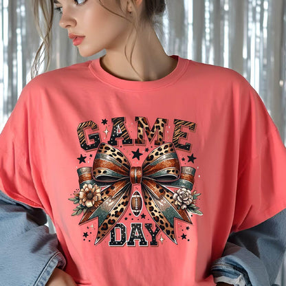May include: A coral-colored t-shirt featuring a graphic print of a large bow with leopard print and glittery textures. The text "GAME DAY" is printed in black with a white outline. The design includes a small football, flowers, and stars.