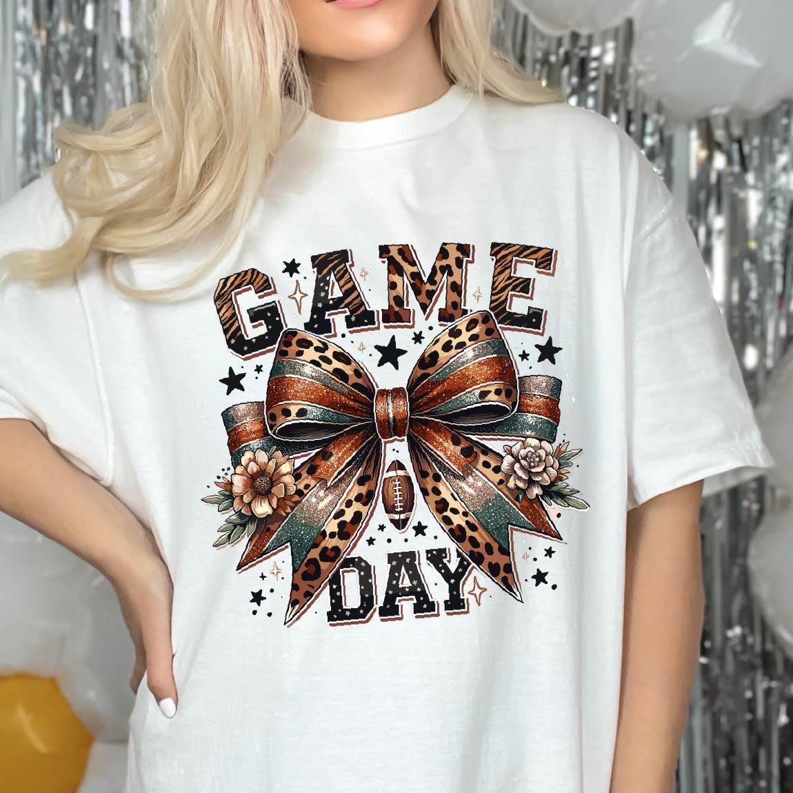 May include: A white t-shirt featuring a graphic print with the text GAME DAY in a leopard print pattern. Centered is a large bow with a leopard print and glittery finish, accented by two flowers and a small football icon, surrounded by stars.