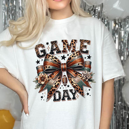 May include: A white t-shirt featuring a graphic print with the text GAME DAY in a leopard print pattern. Centered is a large bow with a leopard print and glittery finish, accented by two flowers and a small football icon, surrounded by stars.