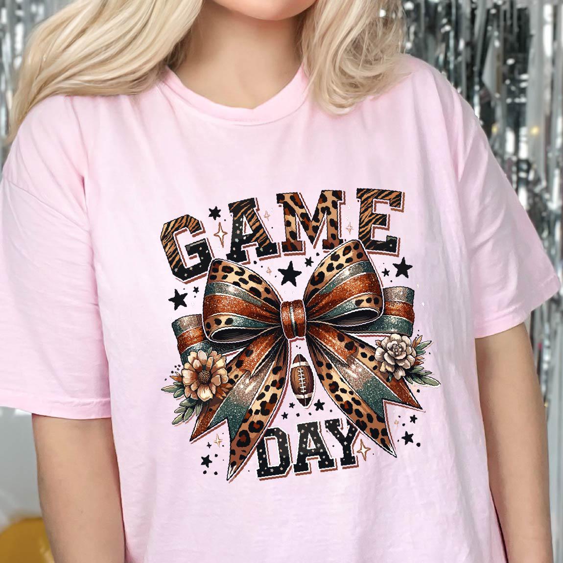 May include: A light pink t-shirt featuring a graphic print with the text "GAME DAY" in a leopard print pattern. The design includes a large central bow with leopard and teal accents, small flowers, stars, and a football icon centered below the bow.