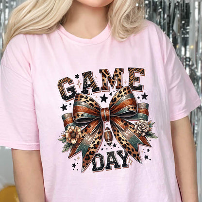 May include: A light pink t-shirt featuring a graphic print with the text "GAME DAY" in a leopard print pattern. The design includes a large central bow with leopard and teal accents, small flowers, stars, and a football icon centered below the bow.