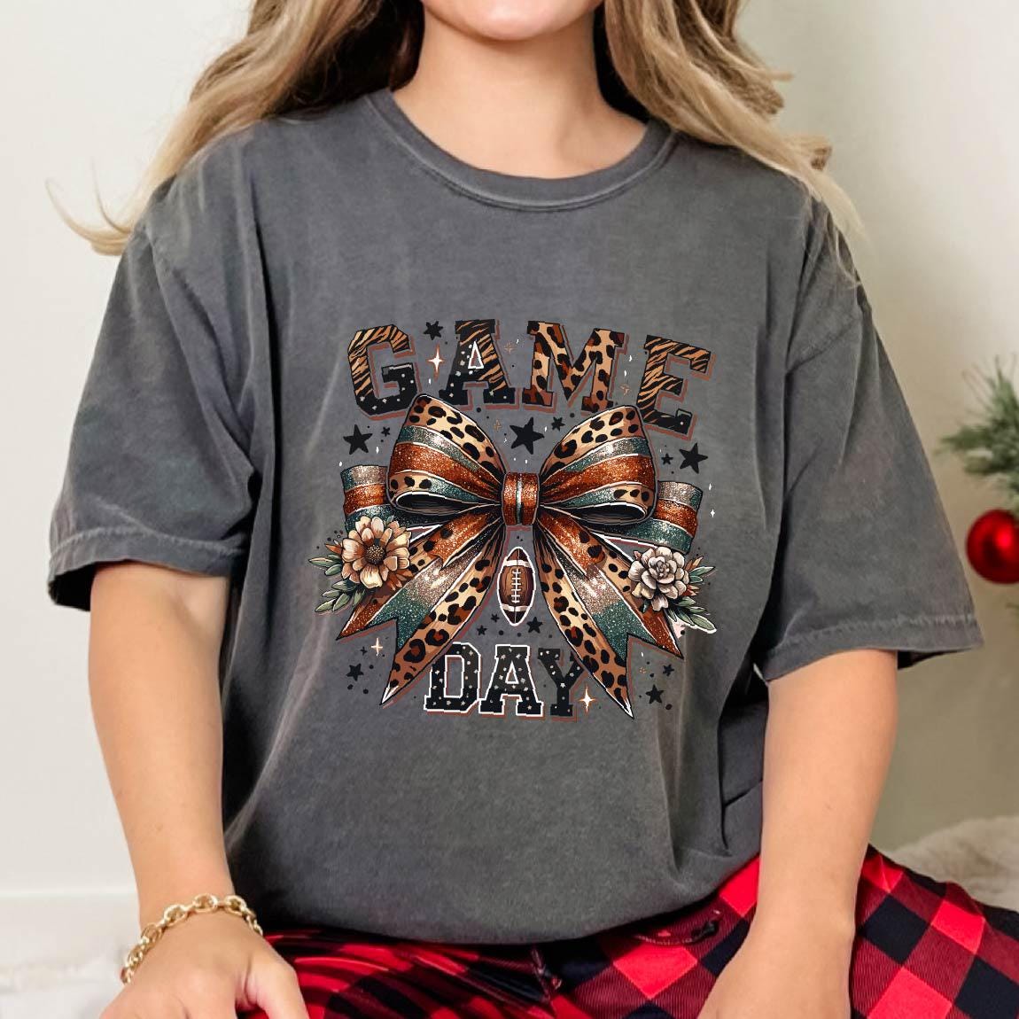 May include: A charcoal gray short-sleeve t-shirt featuring a graphic print with the text GAME DAY. The design includes a large central bow with leopard print, a football icon, and floral accents, surrounded by small black stars on the fabric.