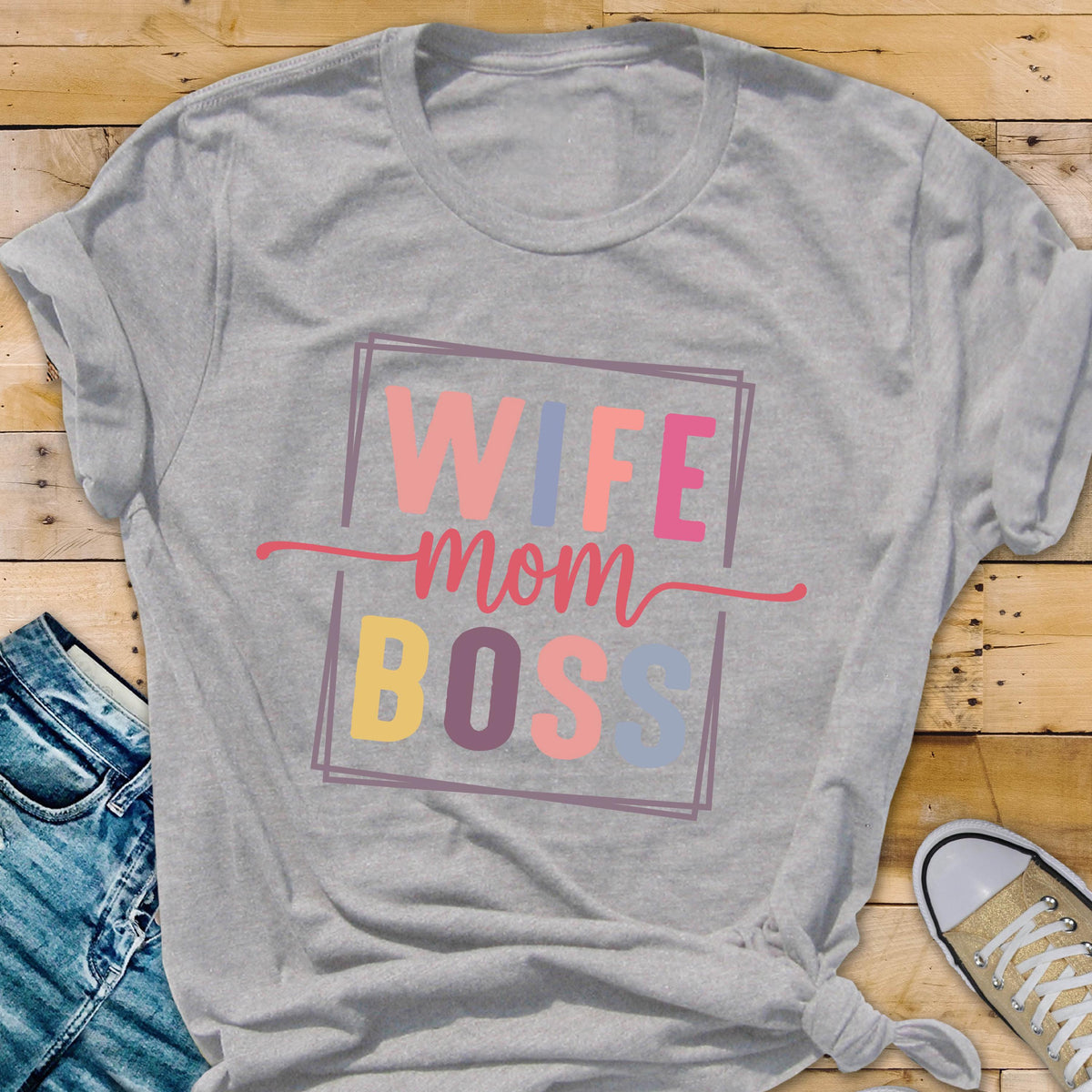 May include: A gray t-shirt with the words "WIFE mom BOSS" in a colorful, rectangular design. The text is in pink, blue, yellow, and peach colors. The shirt is knotted at the bottom and has rolled-up sleeves.
