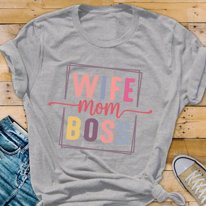 May include: A gray t-shirt with the words "WIFE mom BOSS" in a colorful, rectangular design. The text is in pink, blue, yellow, and peach colors. The shirt is knotted at the bottom and has rolled-up sleeves.