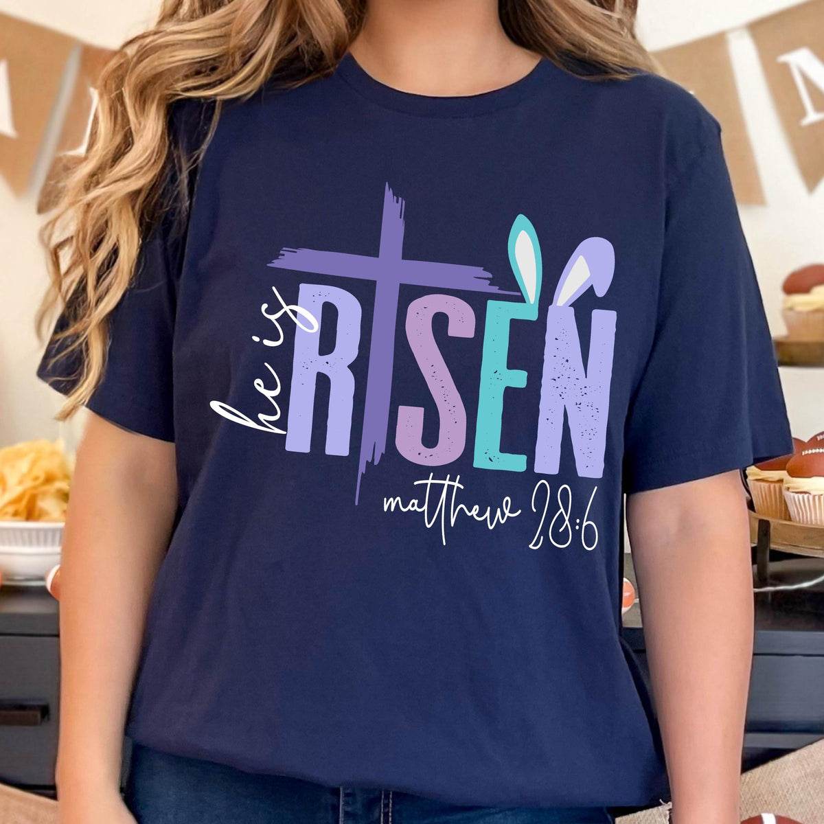 May include: Navy blue t-shirt with a colorful graphic design that says "He is Risen" with a cross and bunny ears. The text "Matthew 28:6" is also included on the shirt.