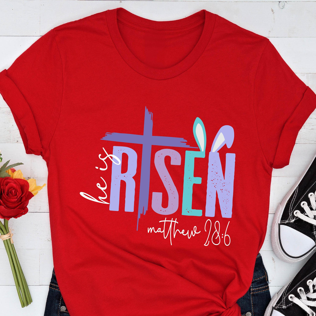 May include: Red t-shirt with a purple, blue, and teal graphic that says "He is Risen" with a cross and bunny ears. The text "matthew 28:6" is below the graphic.