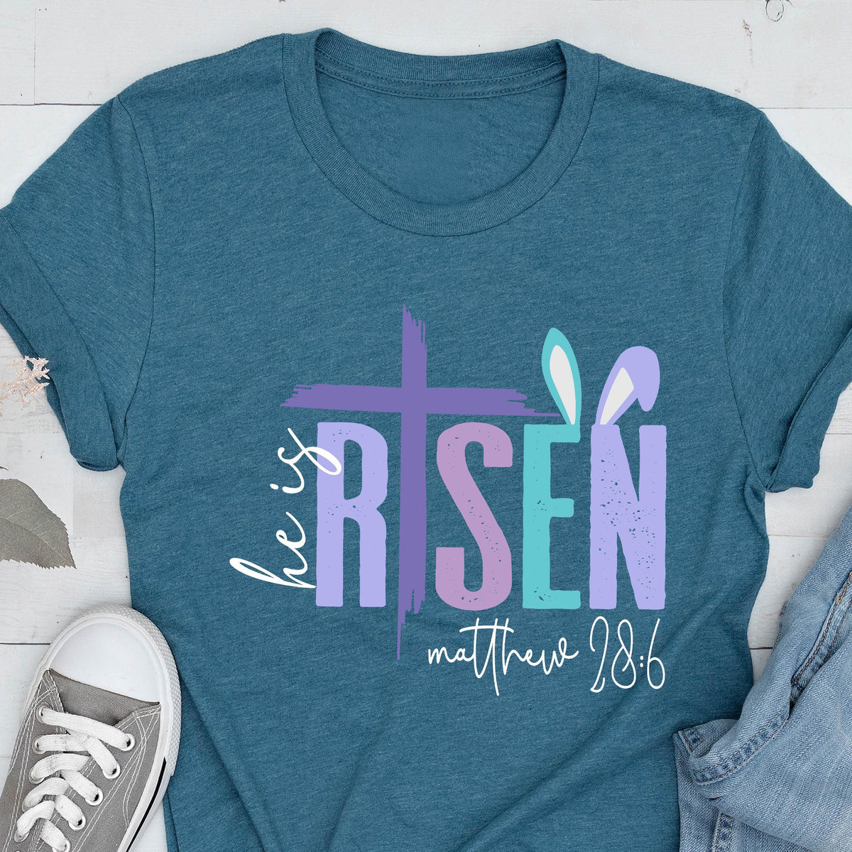 May include: Teal blue t-shirt with a graphic design of a cross and the words "He is Risen" in purple, pink, and blue. The text "Matthew 28:6" is below the graphic.