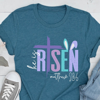 May include: Teal blue t-shirt with a graphic design of a cross and the words "He is Risen" in purple, pink, and blue. The text "Matthew 28:6" is below the graphic.