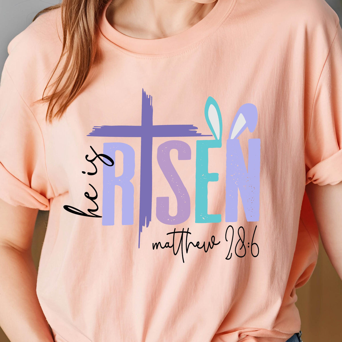 May include: A pink t-shirt with a purple cross and the words "he is risen" in purple, teal, and white. The text "matthew 28:6" is written in black below the words.