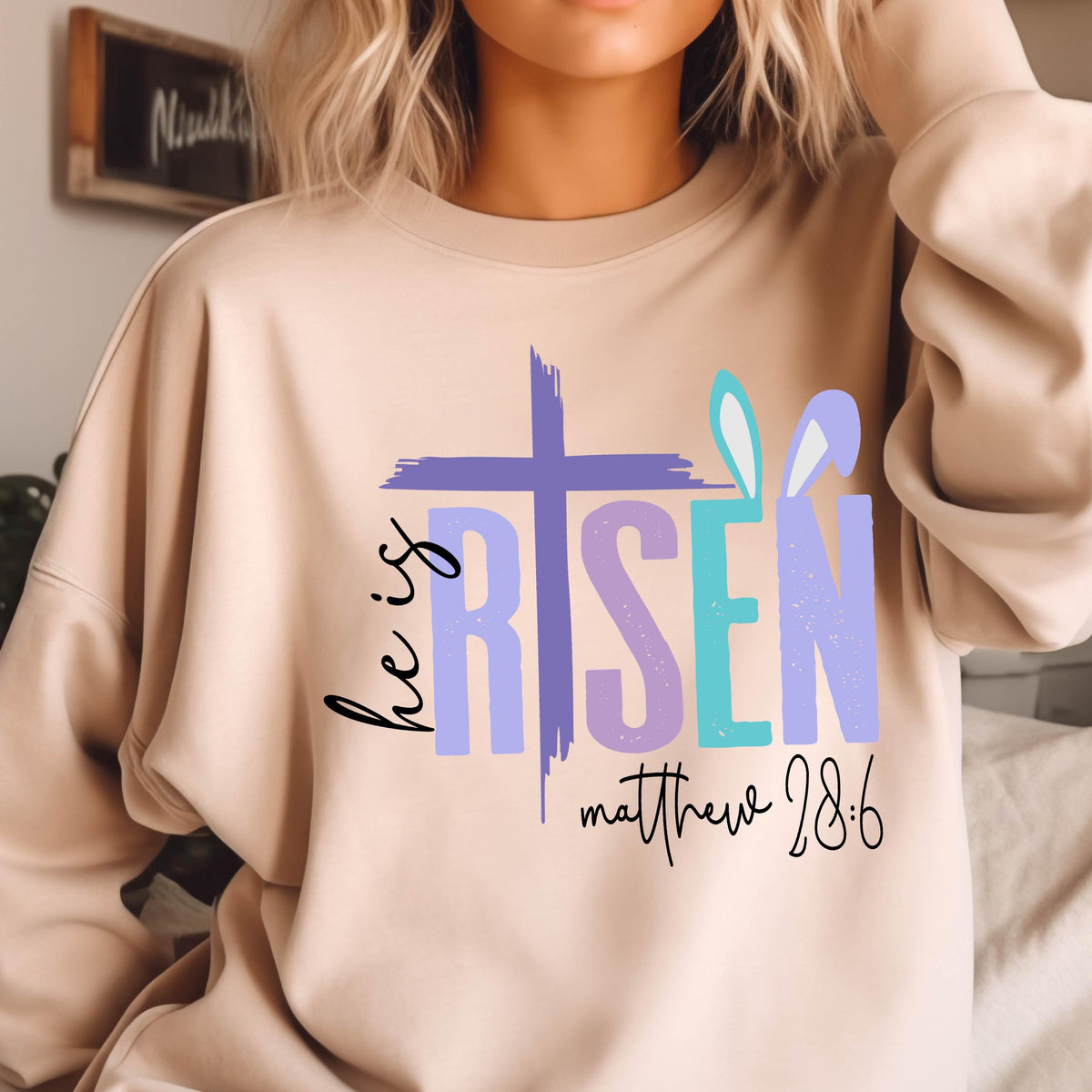 May include: A beige sweatshirt with a graphic design that says "He is Risen" in colorful letters with a cross and a bunny ear design. The text "Matthew 28:6" is printed below the graphic.