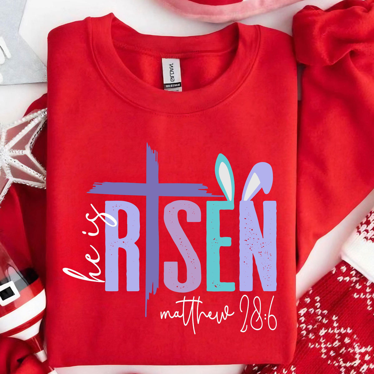 May include: Red sweatshirt with a purple cross and the words "He is Risen" in a rainbow gradient. The text "Matthew 28:6" is written below the graphic.