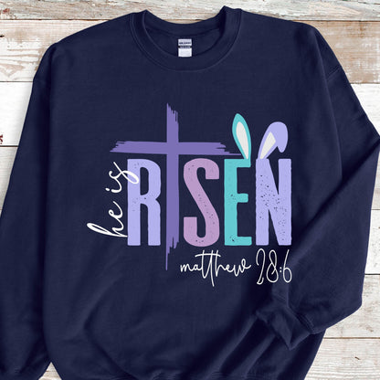 May include: Navy blue sweatshirt with a white graphic design that says "He is Risen" with a purple cross and bunny ears. The text "Matthew 28:6" is also included in the design.