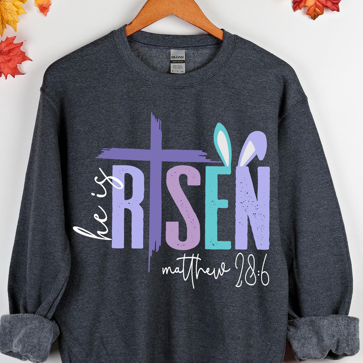 May include: A dark gray sweatshirt with a purple, pink, and blue graphic that says "He is Risen" with bunny ears and the text "Matthew 28:6".