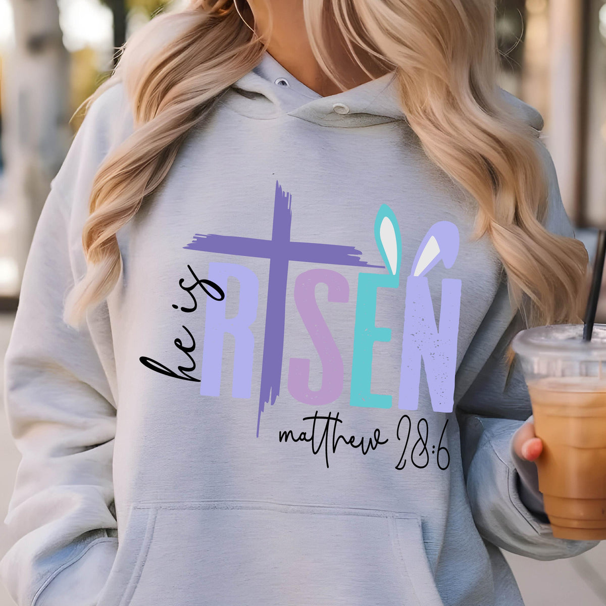 May include: A light gray hoodie with a graphic design that says "He is Risen" in colorful letters with a cross and bunny ears. The text "Matthew 28:6" is printed below the graphic.