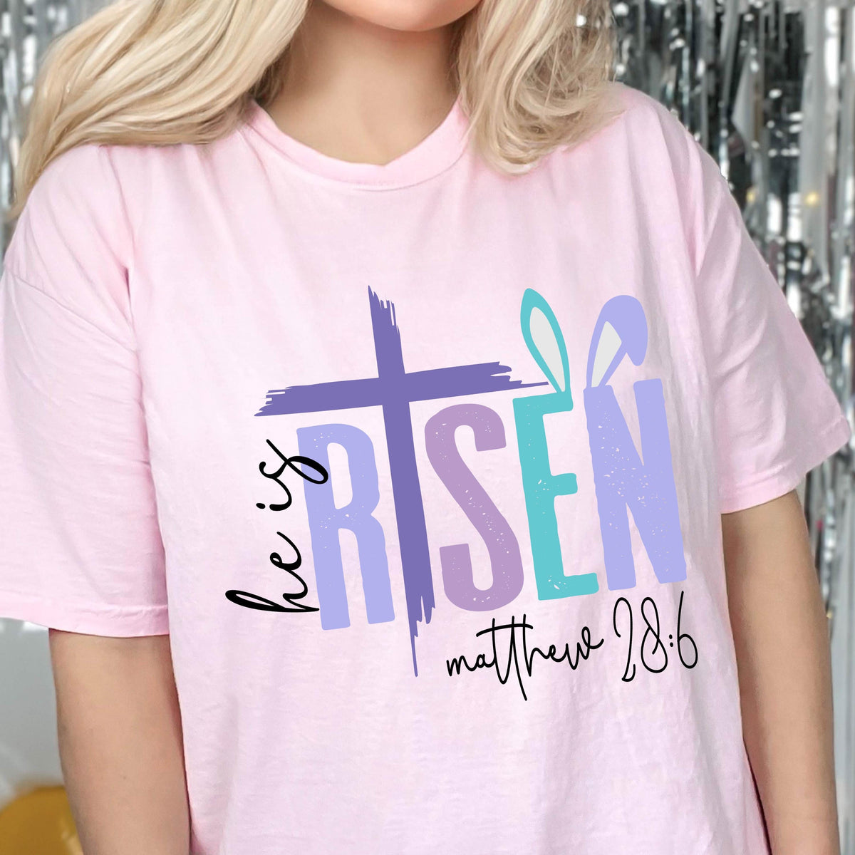 May include: Pink t-shirt with a graphic design that says "He is Risen" with a purple cross and bunny ears. The text "Matthew 28:6" is also included on the shirt.