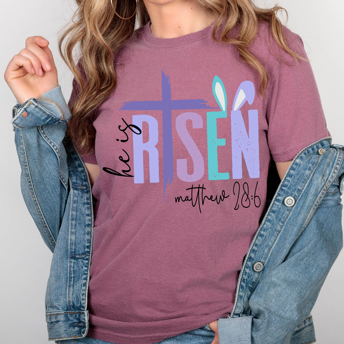May include: A mauve t-shirt with a graphic design of a cross and the words "He is Risen" in colorful letters. The text "Matthew 28:6" is printed below the graphic.