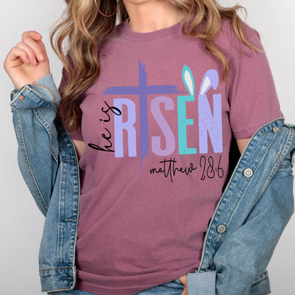 May include: A mauve t-shirt with a graphic design of a cross and the words "He is Risen" in colorful letters. The text "Matthew 28:6" is printed below the graphic.