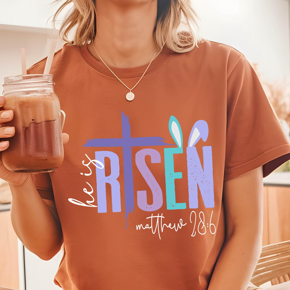 May include: A brown t-shirt with a graphic design that says "He is Risen" with a cross and bunny ears. The text "Matthew 28:6" is also included on the shirt.