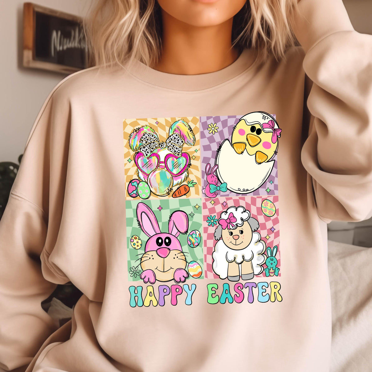 May include: A light pink sweatshirt with a graphic design of four Easter-themed animals in colorful squares. The animals include a bunny wearing sunglasses, a chick in an egg, a bunny with pink ears, and a lamb. The text "Happy Easter" is printed below the animals.