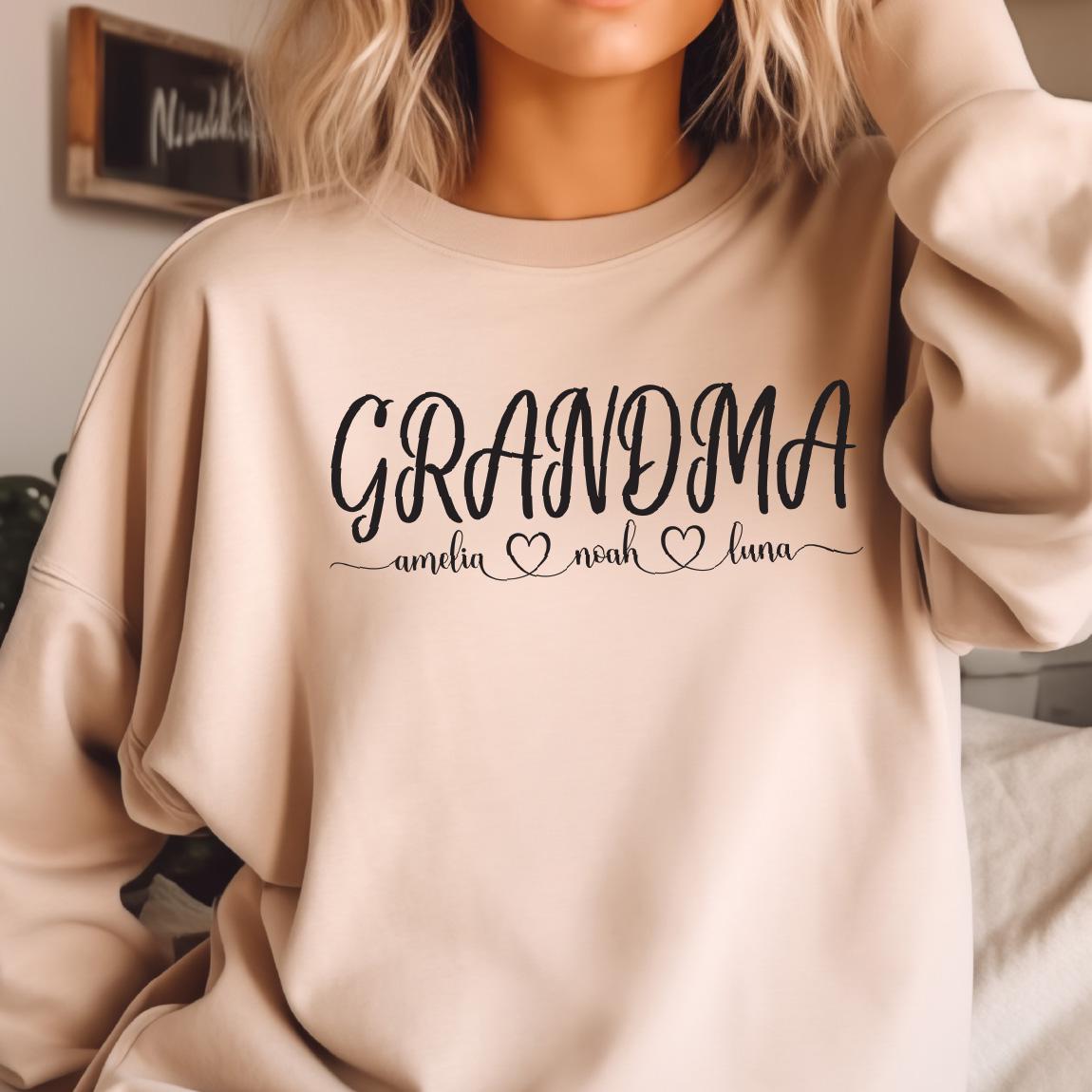 May include: A light beige sweatshirt with the text "GRANDMA" in black cursive lettering. Below the text are three names, "amelia", "noah", and "luna", each with a small heart symbol.