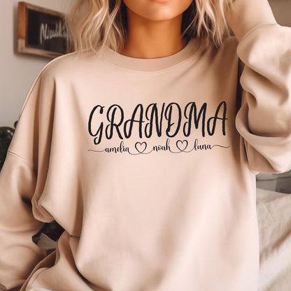 May include: A light beige sweatshirt with the text "GRANDMA" in black cursive lettering. Below the text are three names, "amelia", "noah", and "luna", each with a small heart symbol.