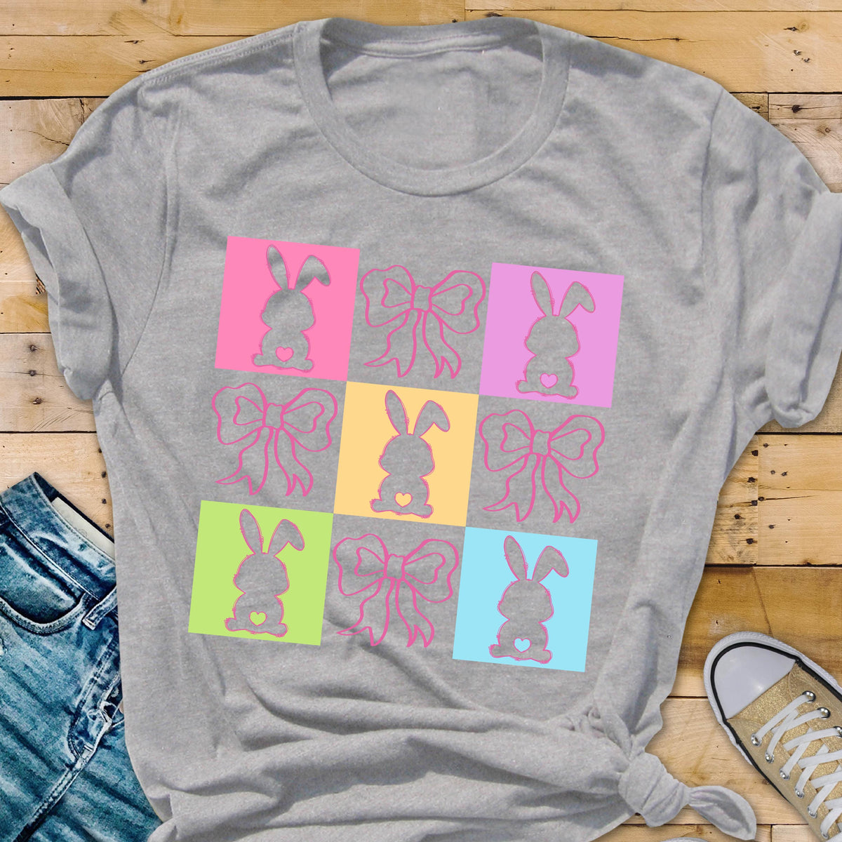 May include: A gray t-shirt with a colorful design of nine squares. Each square features a pink bow and a silhouette of a bunny with a heart in its chest. The squares are arranged in a 3x3 grid.