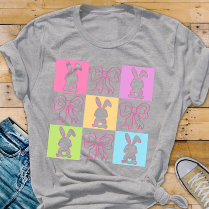 May include: A gray t-shirt with a colorful design of nine squares. Each square features a pink bow and a silhouette of a bunny with a heart in its chest. The squares are arranged in a 3x3 grid.