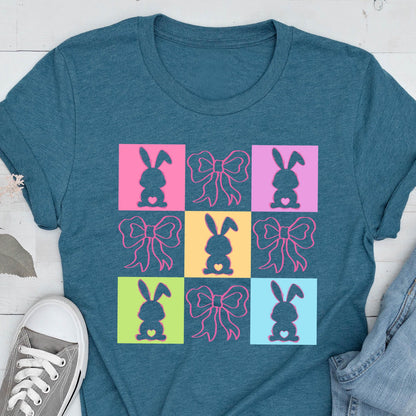 May include: A teal blue t-shirt with a colorful design of nine squares. Each square features a silhouette of a bunny with a heart in its chest and a pink bow. The squares are arranged in a 3x3 grid.