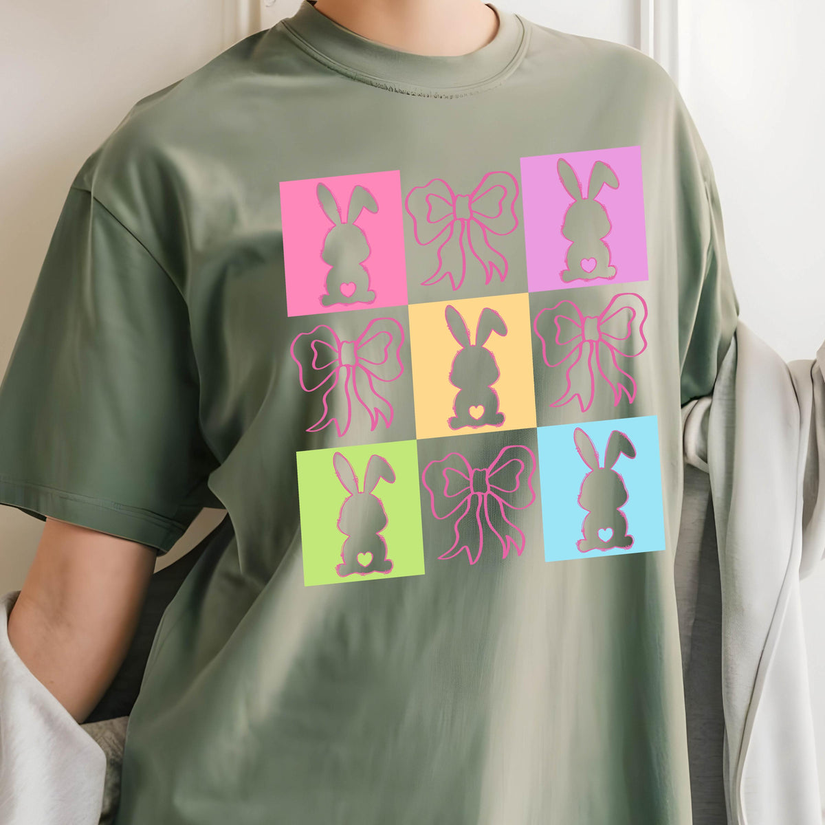 May include: Olive green t-shirt featuring a grid pattern of colorful squares. Each square contains either a bunny silhouette with a heart or a bow. The design is in pink, yellow, blue, and green.