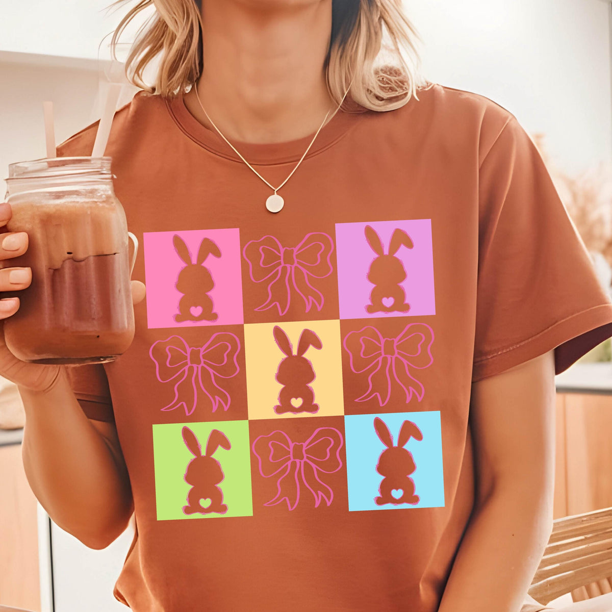 May include: A rust-colored t-shirt featuring a grid pattern of colorful squares. Each square contains either a bunny silhouette or a bow. The bunny silhouettes are in pink, brown, and yellow, while the bows are in pink. The shirt has short sleeves.