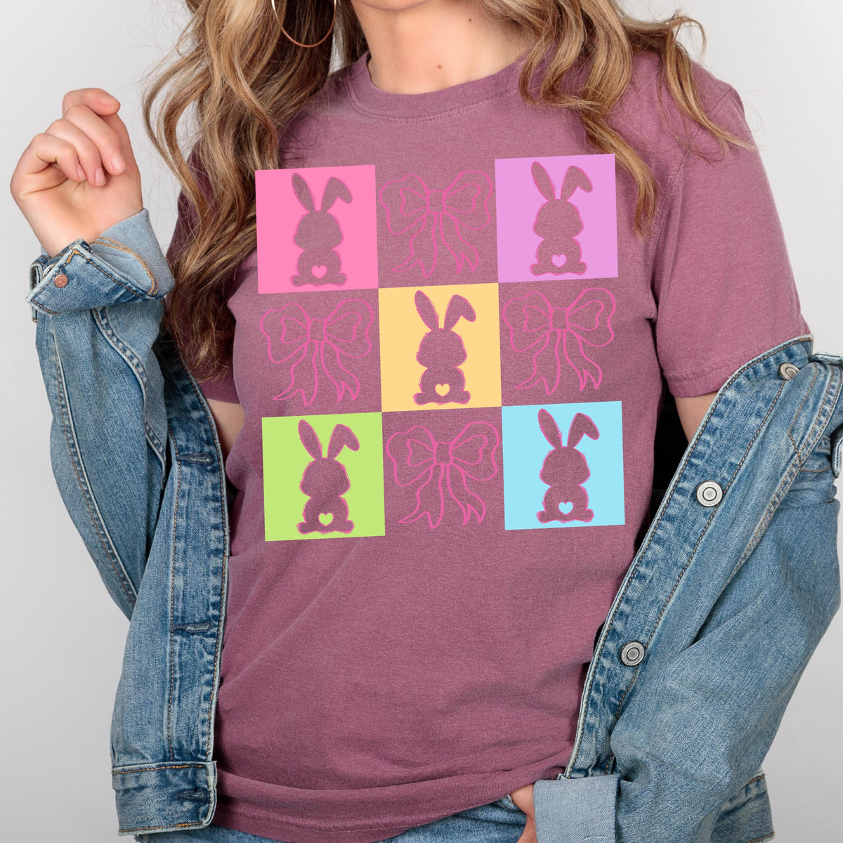May include: A maroon t-shirt featuring a colorful grid pattern with bunny and bow designs. The bunnies are in pink, purple, yellow, green, and blue squares. The bows are in pink. The shirt has short sleeves and a crew neck.
