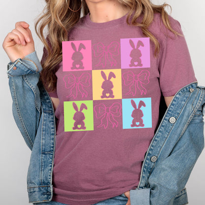May include: A maroon t-shirt featuring a colorful grid pattern with bunny and bow designs. The bunnies are in pink, purple, yellow, green, and blue squares. The bows are in pink. The shirt has short sleeves and a crew neck.
