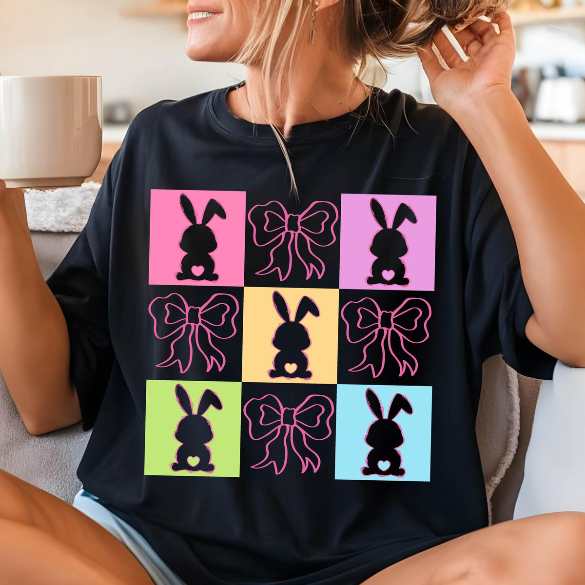 May include: Black t-shirt featuring a grid design with Easter-themed graphics. The design includes bunny silhouettes with hearts and pink bows set against colorful squares of pink, yellow, green, and blue. A casual, festive top.