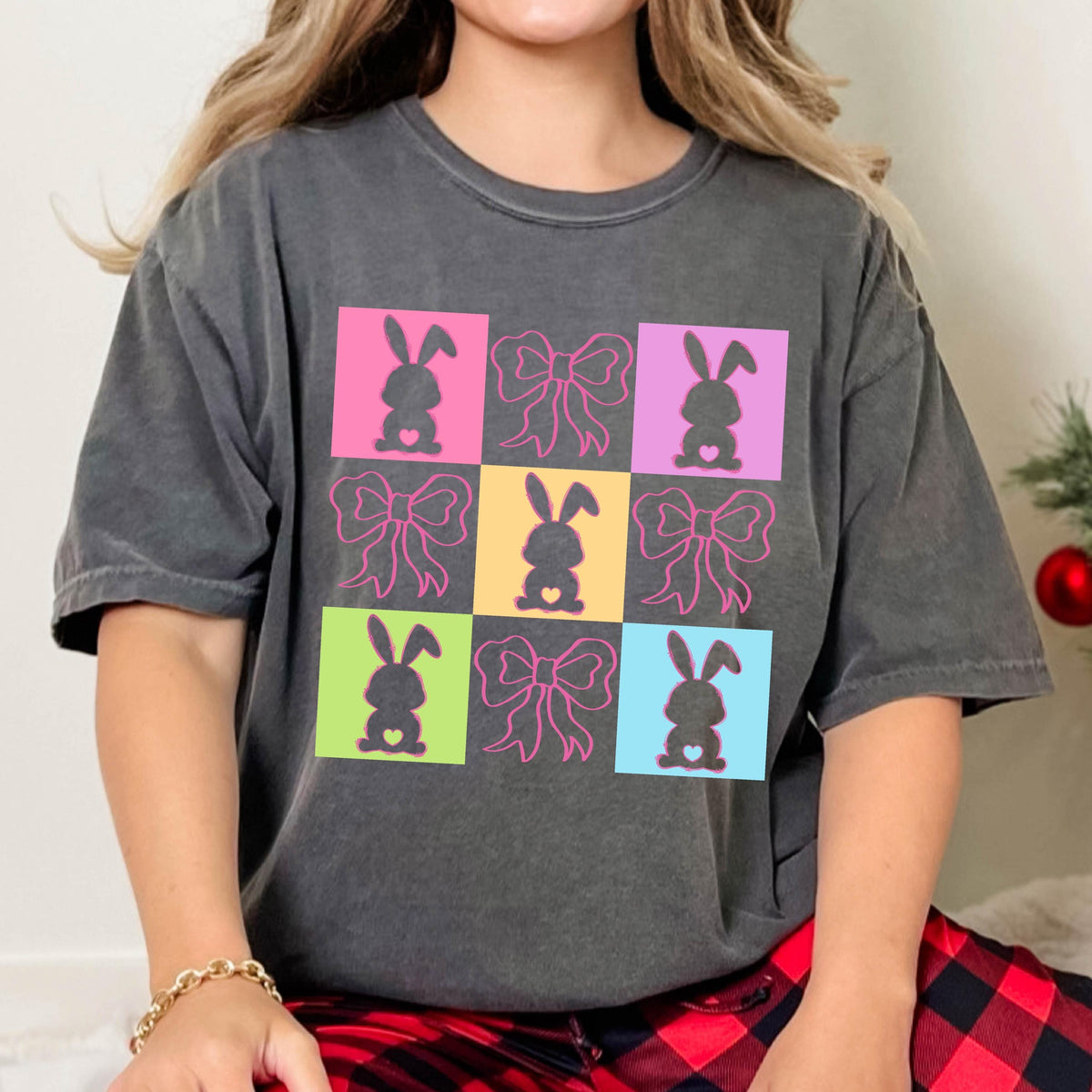 May include: Dark gray t-shirt featuring a colorful grid pattern with Easter-themed designs. The design includes bunny silhouettes and bows in pink, yellow, green, and blue squares. The shirt has a relaxed fit.