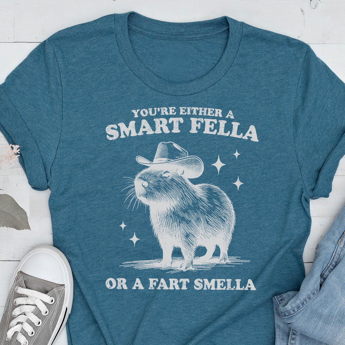 May include: Teal t-shirt with a white graphic of a capybara wearing a cowboy hat. The text reads "YOU'RE EITHER A SMART FELLA OR A FART SMELLA". The shirt is made of a soft material and is suitable for casual wear.