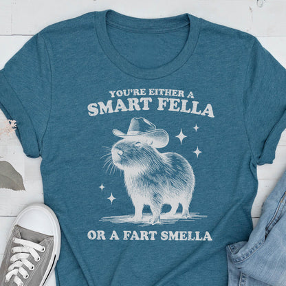 May include: Teal t-shirt with a white graphic of a capybara wearing a cowboy hat. The text reads "YOU'RE EITHER A SMART FELLA OR A FART SMELLA". The shirt is made of a soft material and is suitable for casual wear.
