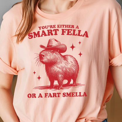 May include: A light peach-colored t-shirt featuring a red illustration of a capybara wearing a cowboy hat. The text above reads "YOU'RE EITHER A SMART FELLA" and below "OR A FART SMELLA". Four red star shapes are also present.