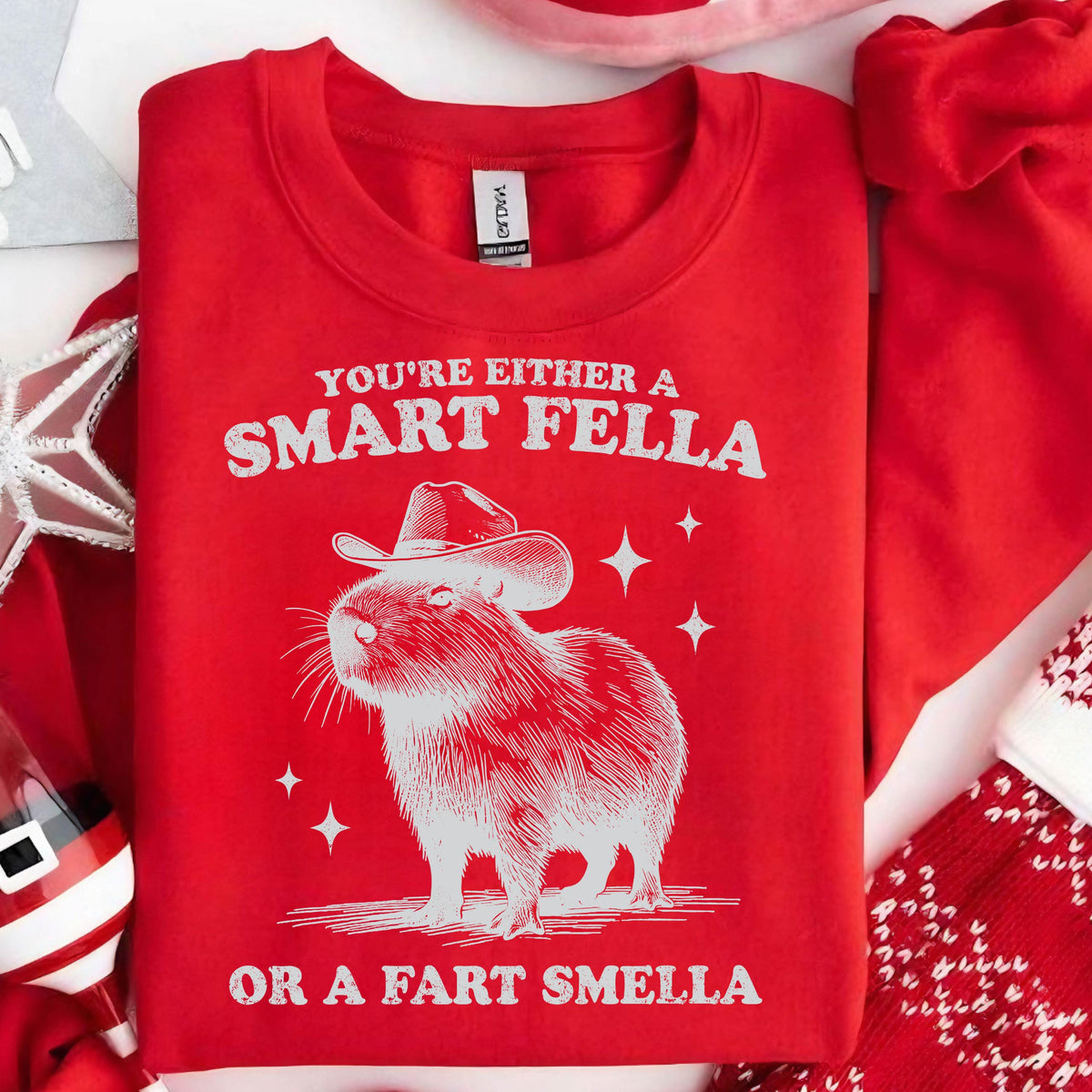 May include: Red sweatshirt with a white graphic of a capybara wearing a cowboy hat. The text reads "YOU'RE EITHER A SMART FELLA OR A FART SMELLA". The sweatshirt is folded and surrounded by Christmas decorations.