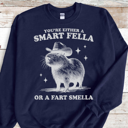 May include: Navy blue sweatshirt with a graphic of a capybara wearing a cowboy hat. The text reads "YOU'RE EITHER A SMART FELLA OR A FART SMELLA" in white. The design includes star accents.
