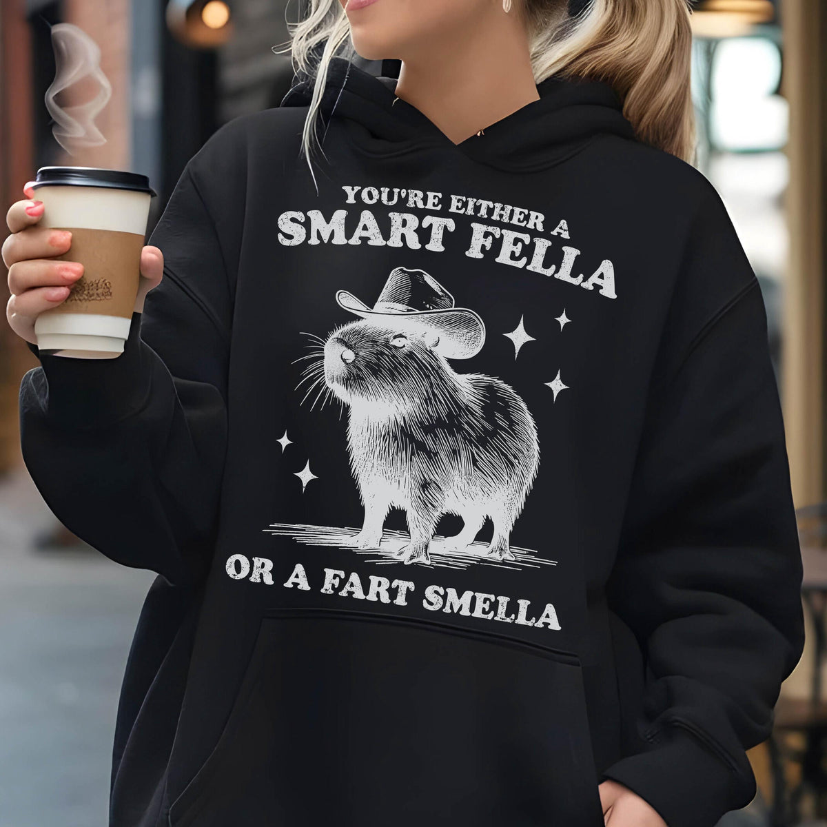 May include: Black hoodie with a graphic of a capybara wearing a cowboy hat. The text reads "You're either a Smart Fella or a Fart Smella" in white. The design includes stars.