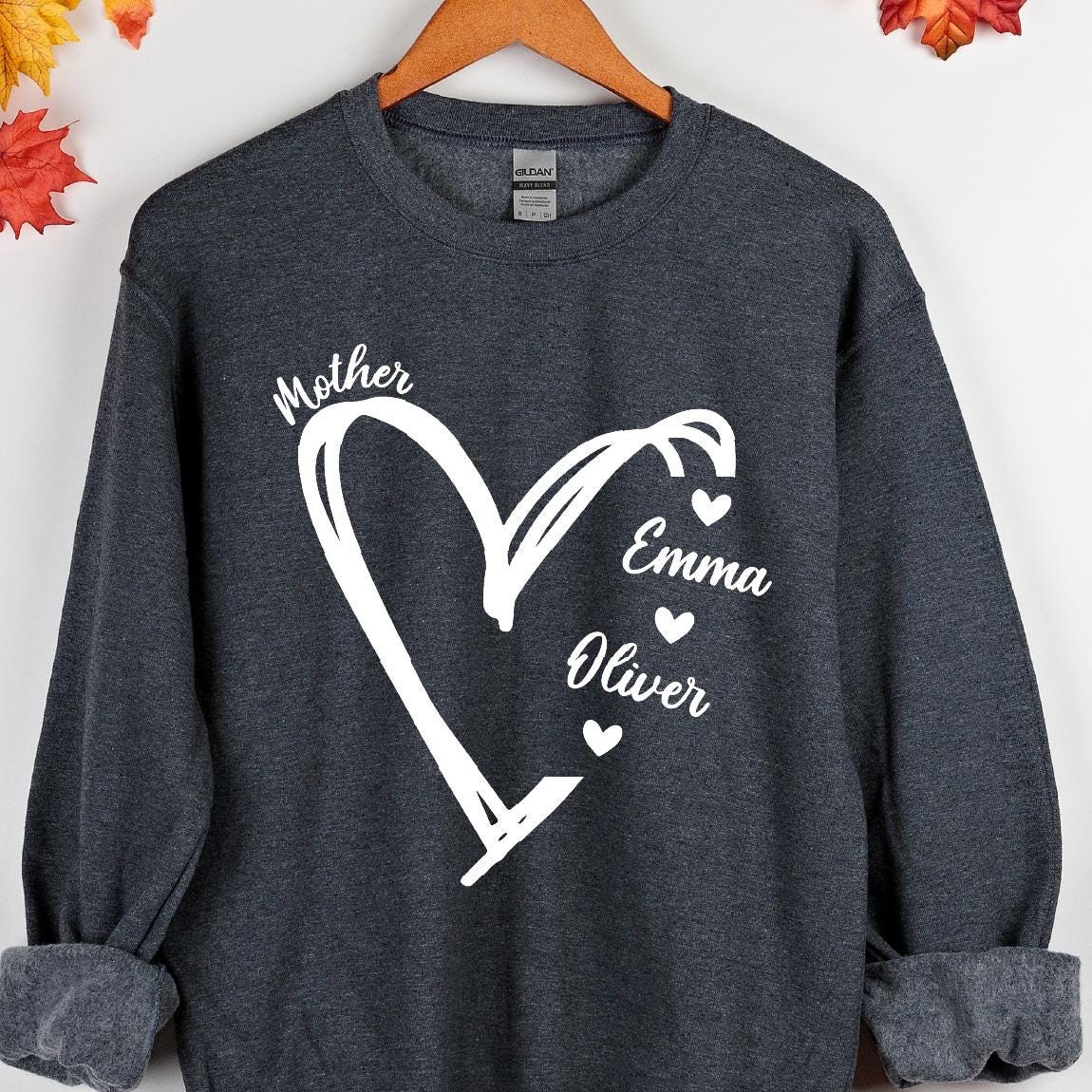 May include: A dark gray sweatshirt with a white heart design. The heart has the word "Mother" at the top and the names "Emma" and "Oliver" inside the heart.