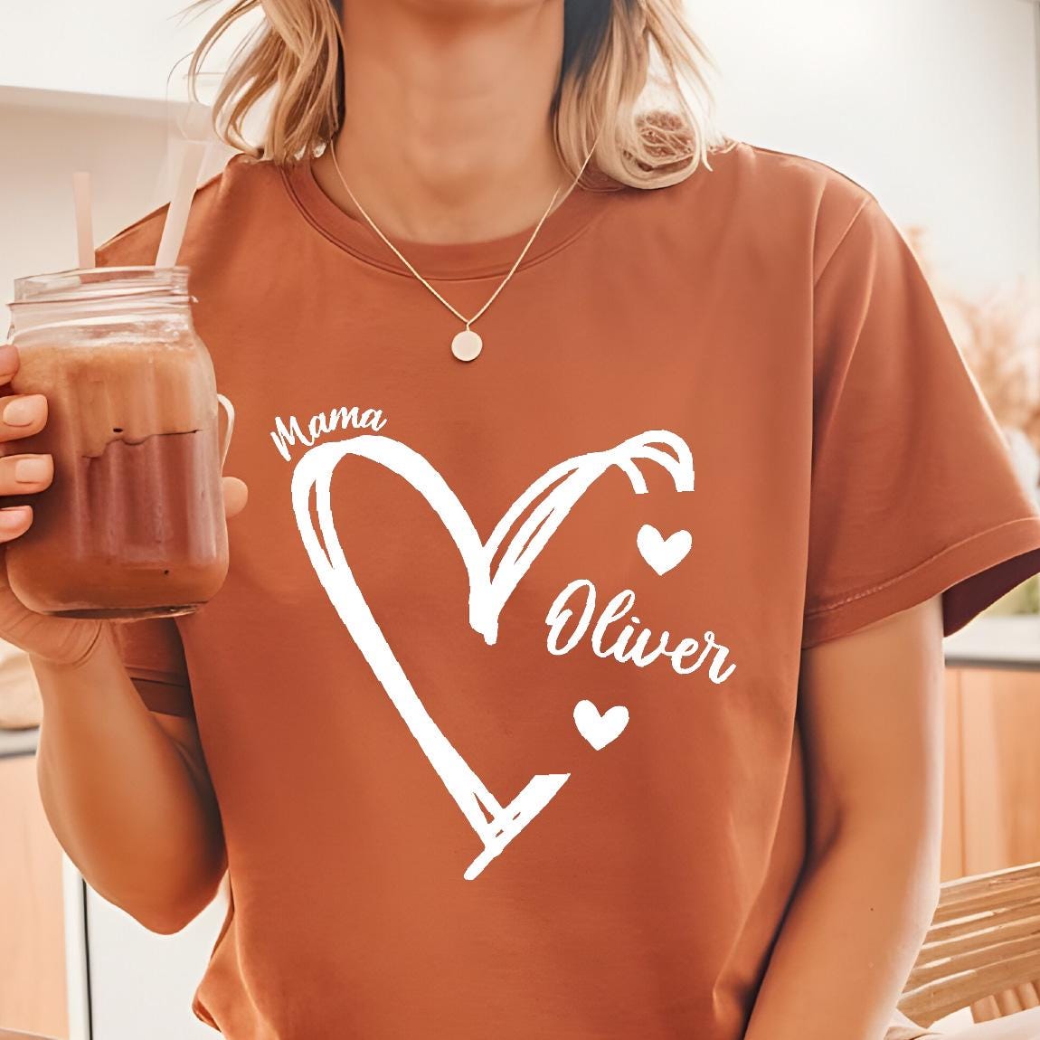 May include: A rust-colored short-sleeve t-shirt featuring a white graphic print of a hand-drawn heart outline. The text "Mama" is positioned above the heart, while the name "Oliver" is written inside the heart with two small hearts.