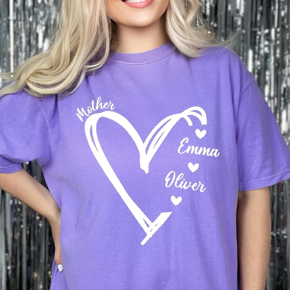 May include: A light purple short-sleeve t-shirt featuring a large white hand-drawn heart outline. The text "Mother" is printed above the heart, while the names "Emma" and "Oliver" are printed to the right, each accompanied by a small heart icon.