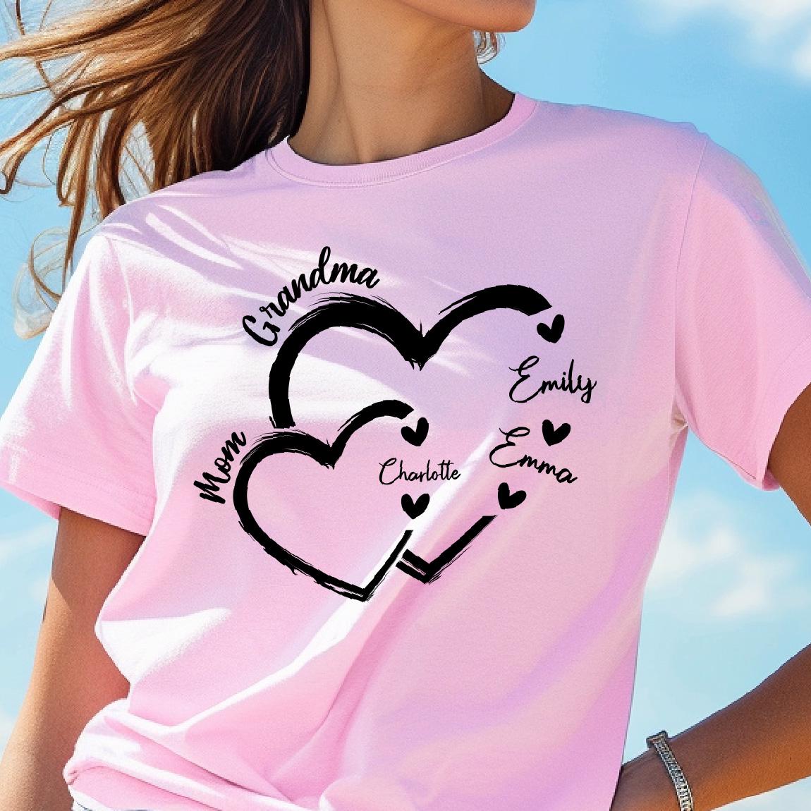 May include: Pink t-shirt with a black design of two interlocking hearts. The text "Grandma" is above the hearts, and the text "Mom" is to the left of the hearts. Inside the hearts are the names "Emily", "Emma", and "Charlotte".